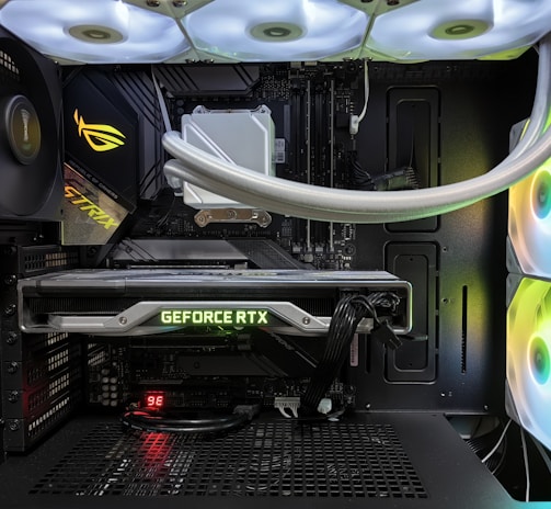 Inside view of a computer case showcasing a sophisticated gaming setup. Visible components include a GeForce RTX graphics card illuminated with green lighting, an ASUS motherboard, cooling fans with LED lighting, and a liquid cooling system with white tubes. Cables are neatly organized, and overall, the hardware suggests a high-performance machine.