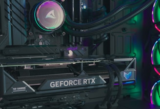 The image depicts the inside of a computer case featuring a high-performance graphics card labeled 'GEFORCE RTX' with RGB lighting. The cooling fans are illuminated with vibrant colors, creating a visually striking look. Various cables and components are neatly arranged, highlighting advanced hardware and a focus on aesthetics.